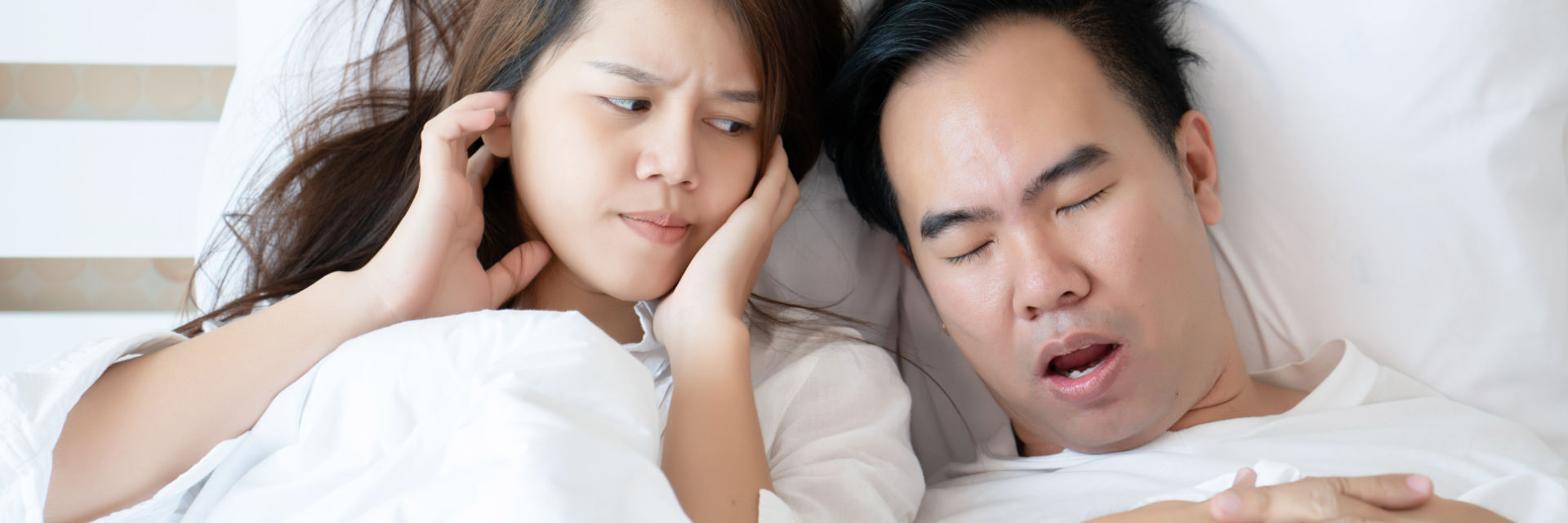 A snoring man sleeping next to an annoyed woman blocking her ears.